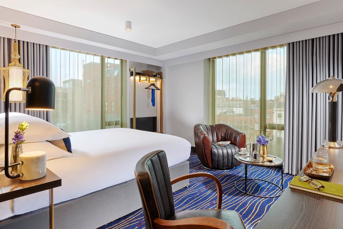 A modern hotel room with a large bed, a leather armchair, a small table with a tray, and a desk with a lamp and vase.