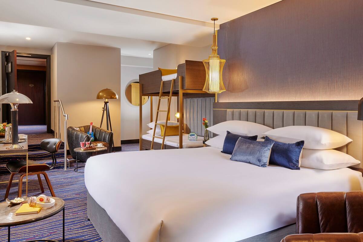 A spacious hotel room with a large bed, a desk with a lamp, a chair, and a bedside table with a vase of flowers.