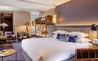 Quarto de hotel espaçoso, com uma cama grande, uma escrivaninha com luminária, uma cadeira e uma mesinha de cabeceira com um vaso de flores.