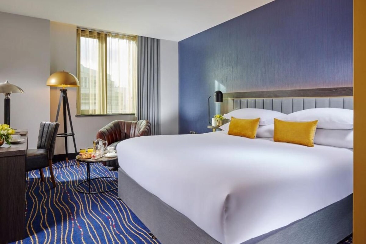A hotel room with a large bed, a desk with a lamp, a chair, and a window with curtains.