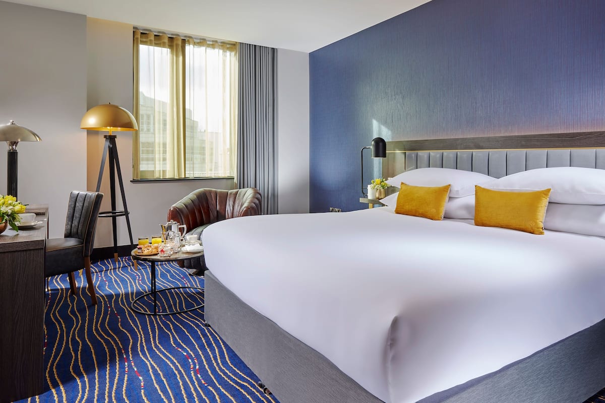 A hotel room with a large bed, a bedside table with a lamp, a chair, a desk with a chair, and a window with curtains.