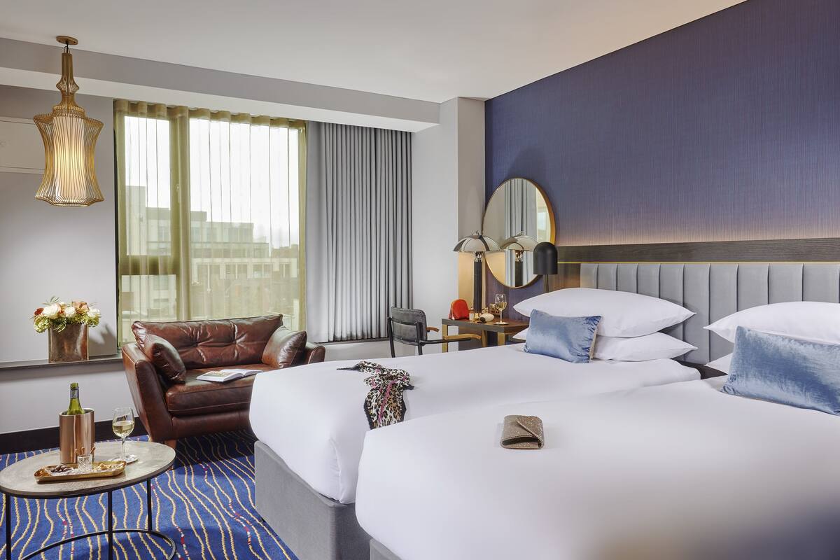 A hotel room with a large bed, a leather armchair, a small table with a bottle and glasses, and a view of the city through the window.