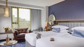 A hotel room with a large bed, a leather armchair, a small table with a bottle and glasses, and a view of the city through the window.