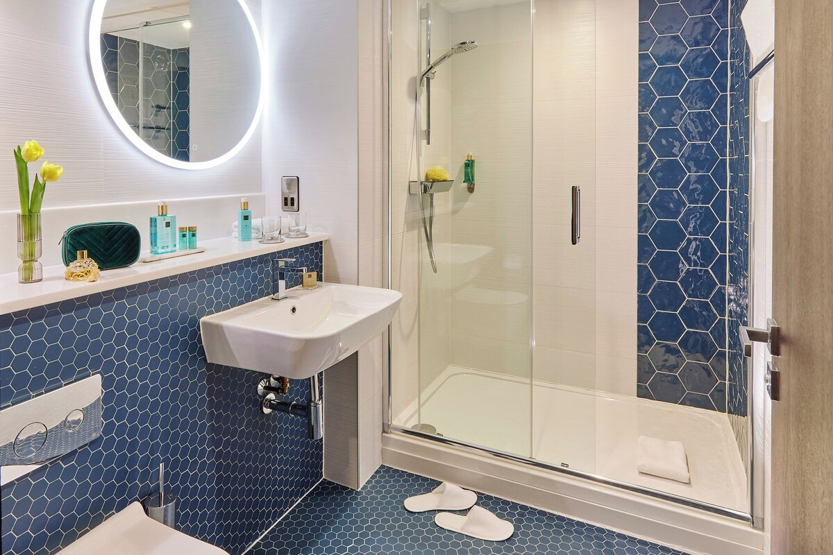 A modern bathroom with a glass-enclosed shower, a white sink, and a mirror with a circular light.