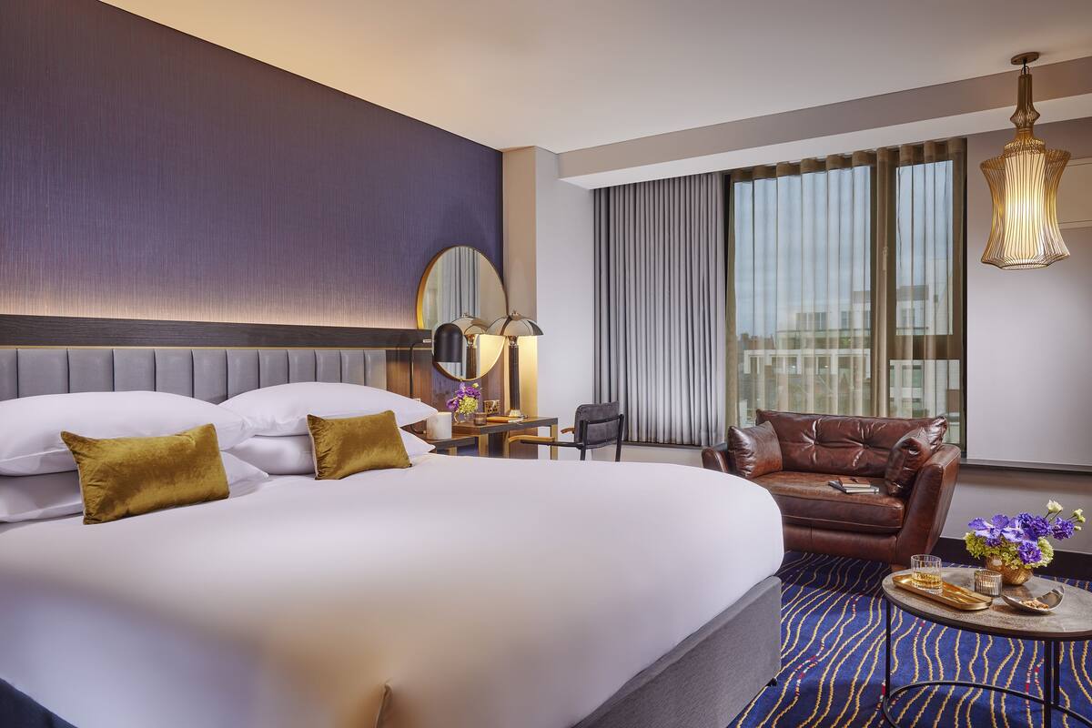 A hotel room with a large bed, a leather armchair, a small table with a tray, and a large window with curtains.