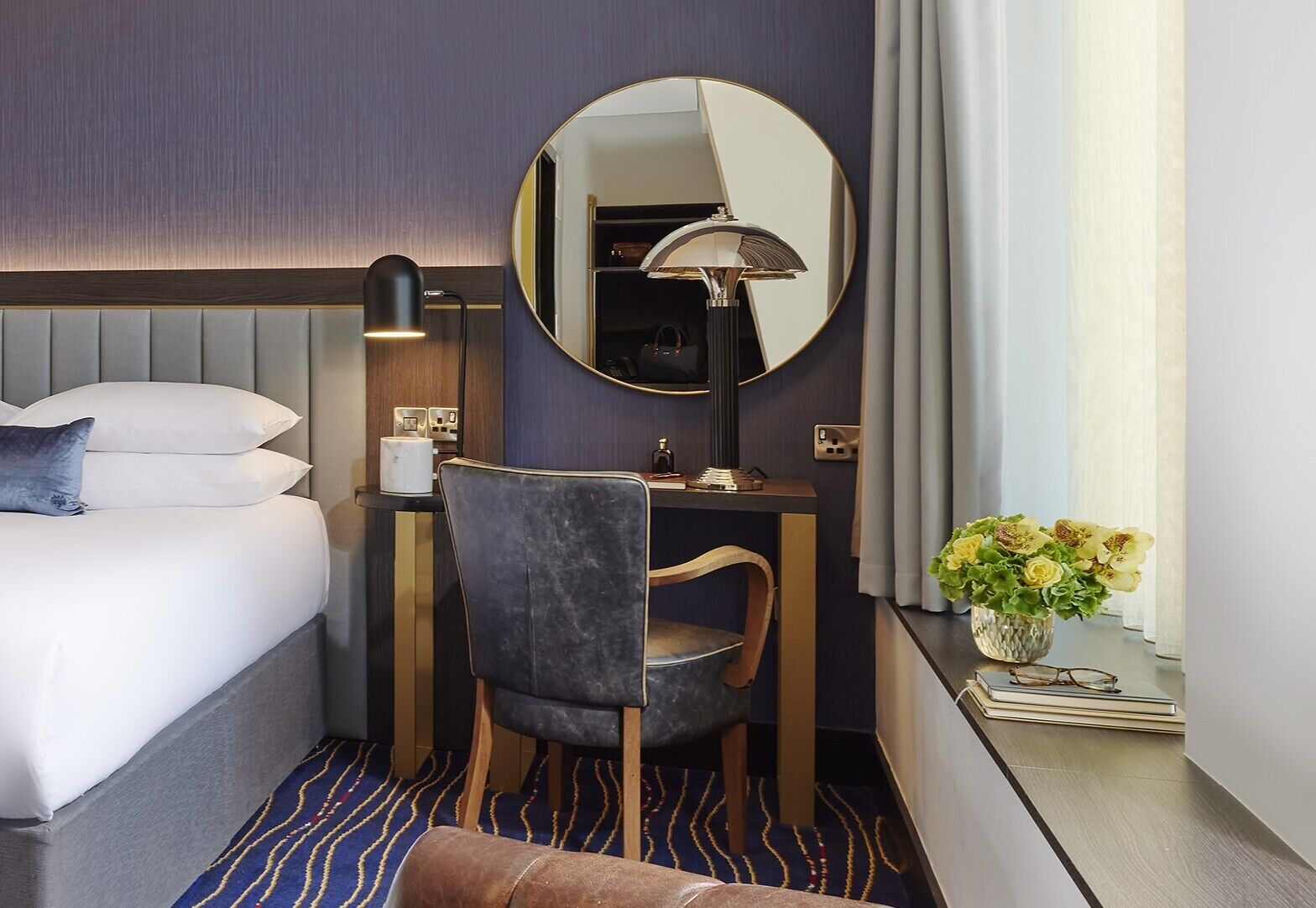A hotel room with a bed, a desk with a chair, a round mirror, a lamp, and a vase with flowers.
