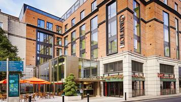 A modern hotel with a glass facade, outdoor seating area, and a visible entrance.