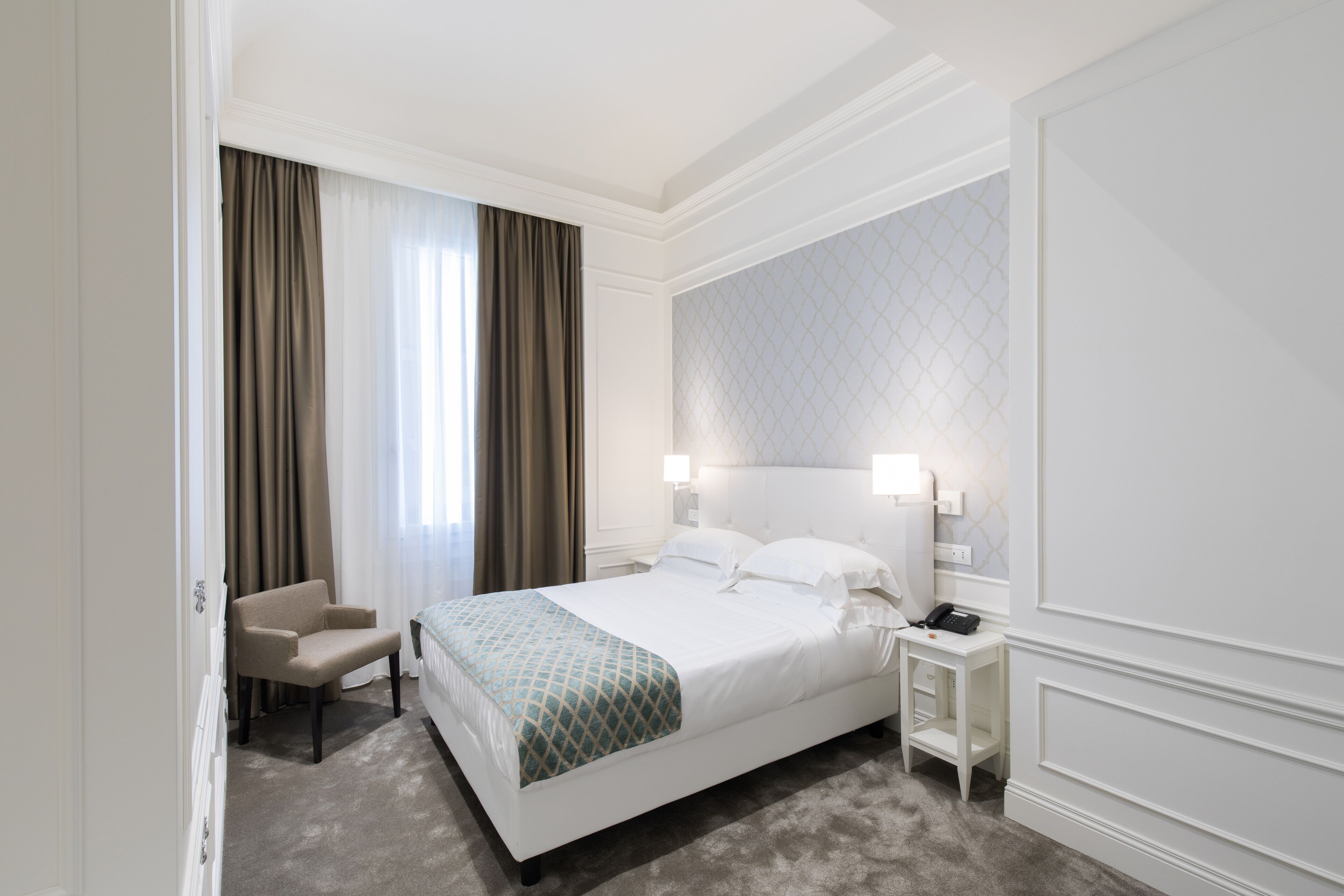 Camera d'albergo con un letto grande, una sedia, un comodino con il telefono e una finestra con tende.