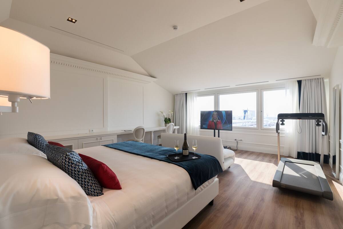 A modern hotel room with a large bed, a treadmill, a desk with a chair, and a TV.