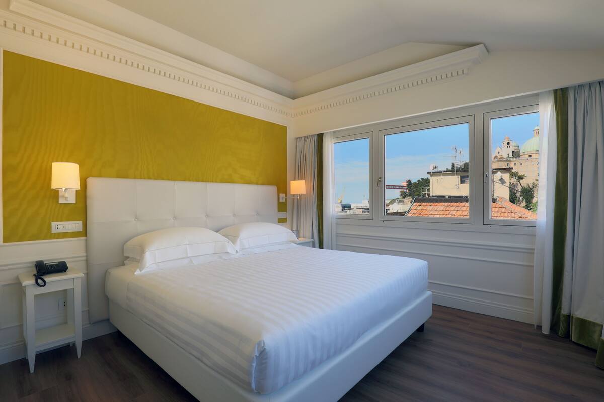 A hotel room with a large bed, a bedside table with a lamp, and a window with a city view.