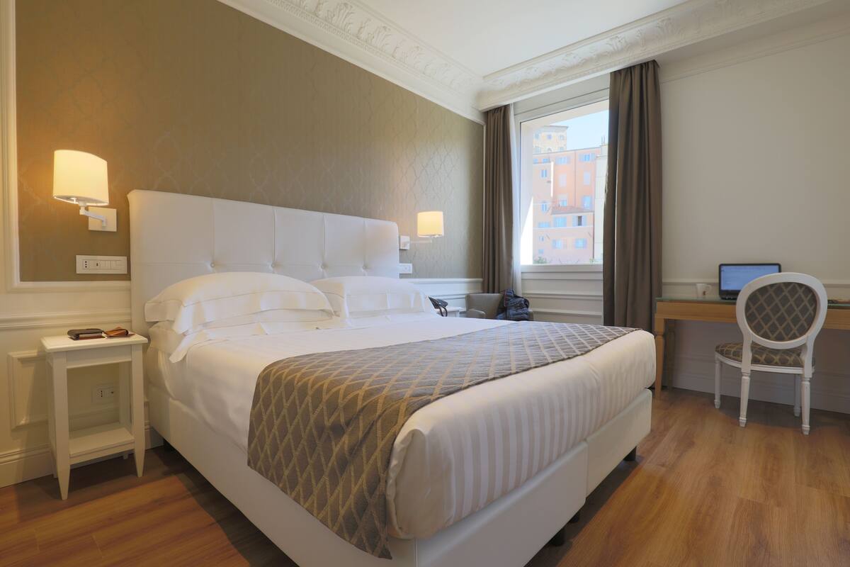 A hotel room with a large bed, bedside tables, a desk with a chair, and a window with curtains.