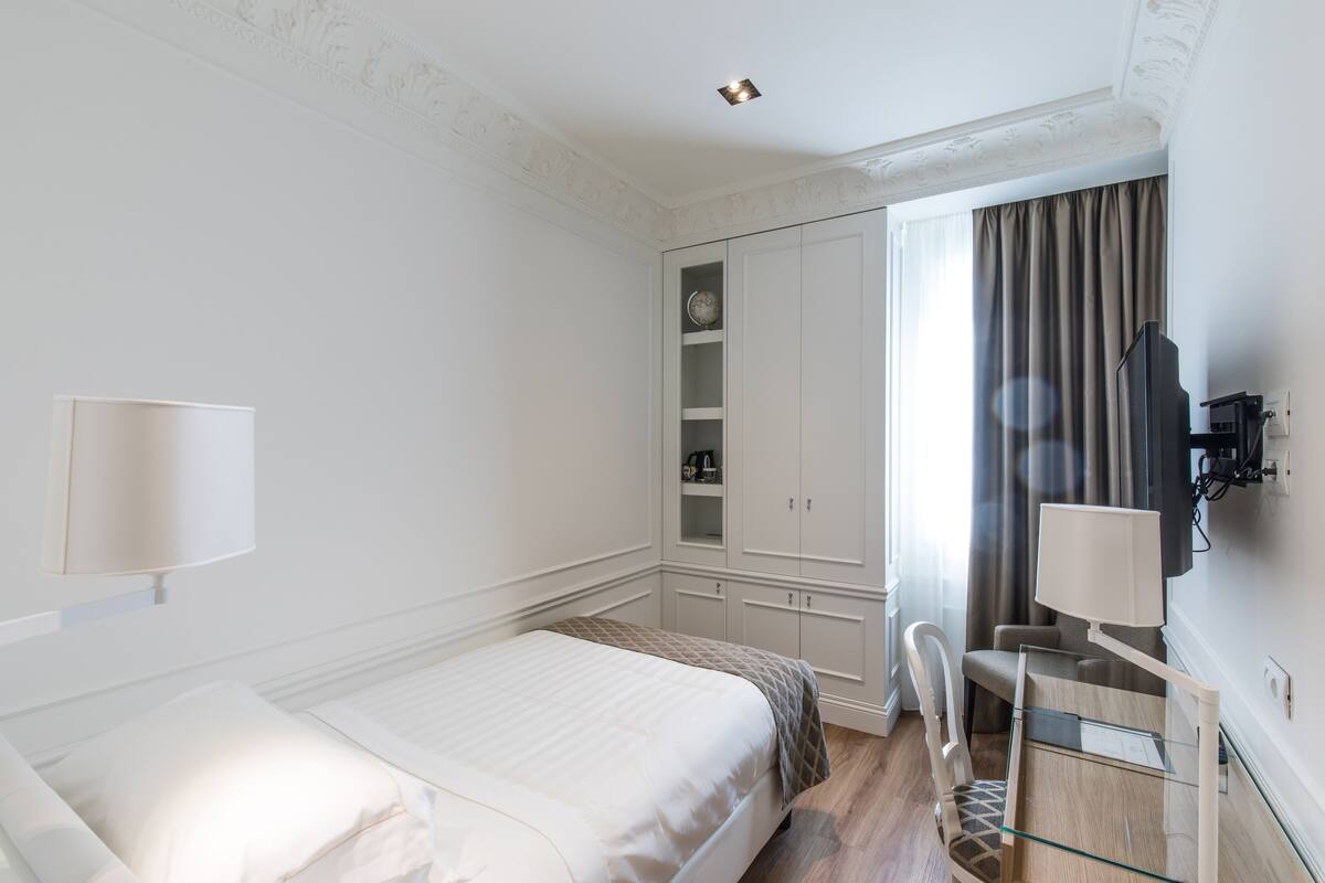 A hotel room with a bed, a desk with a chair, a TV, a wardrobe, and a window with curtains.