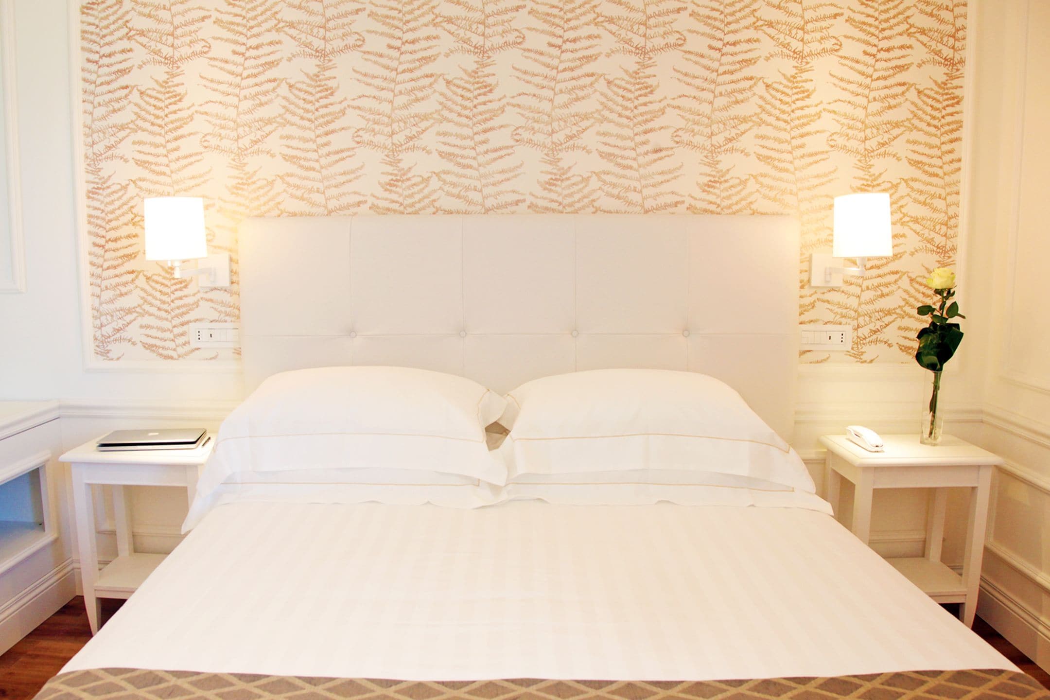 A neatly made bed with a white headboard, two bedside tables with lamps, and a patterned wallpaper.