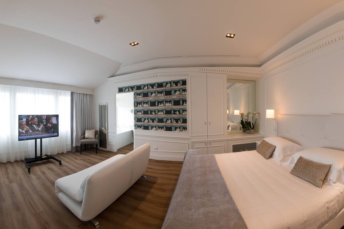 A hotel room with a large bed, a TV, a seating area, and a minibar.