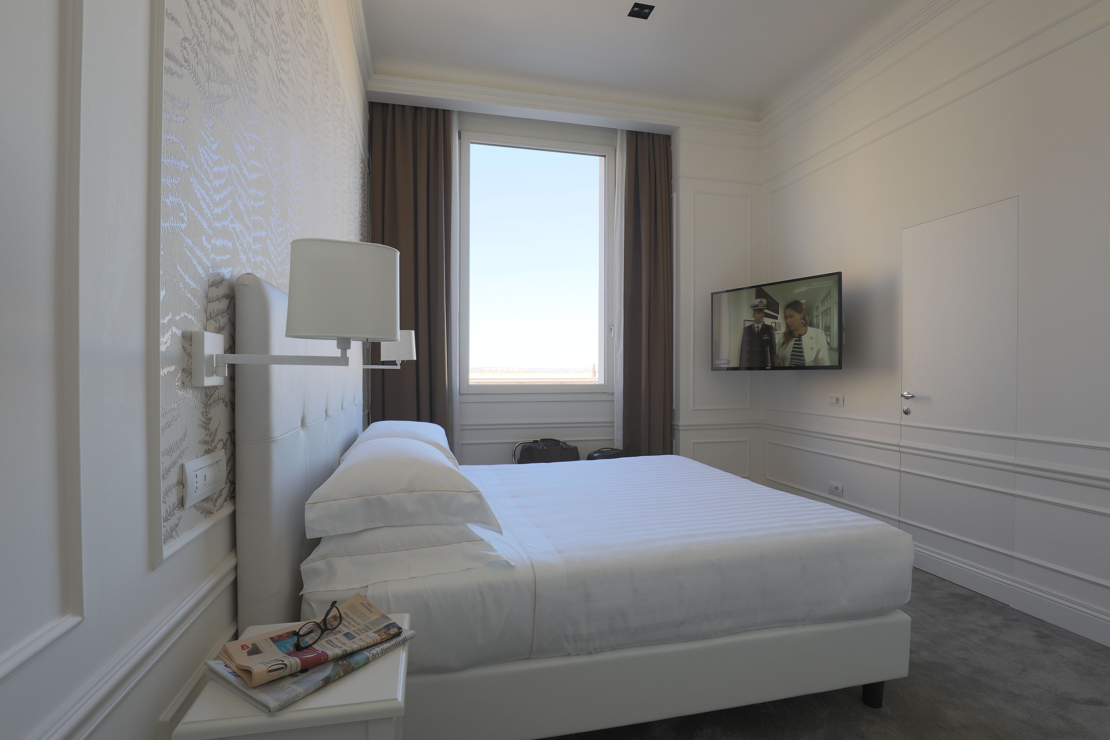 A hotel room with a neatly made bed, a bedside table with a lamp, a window with curtains, and a television on the wall.