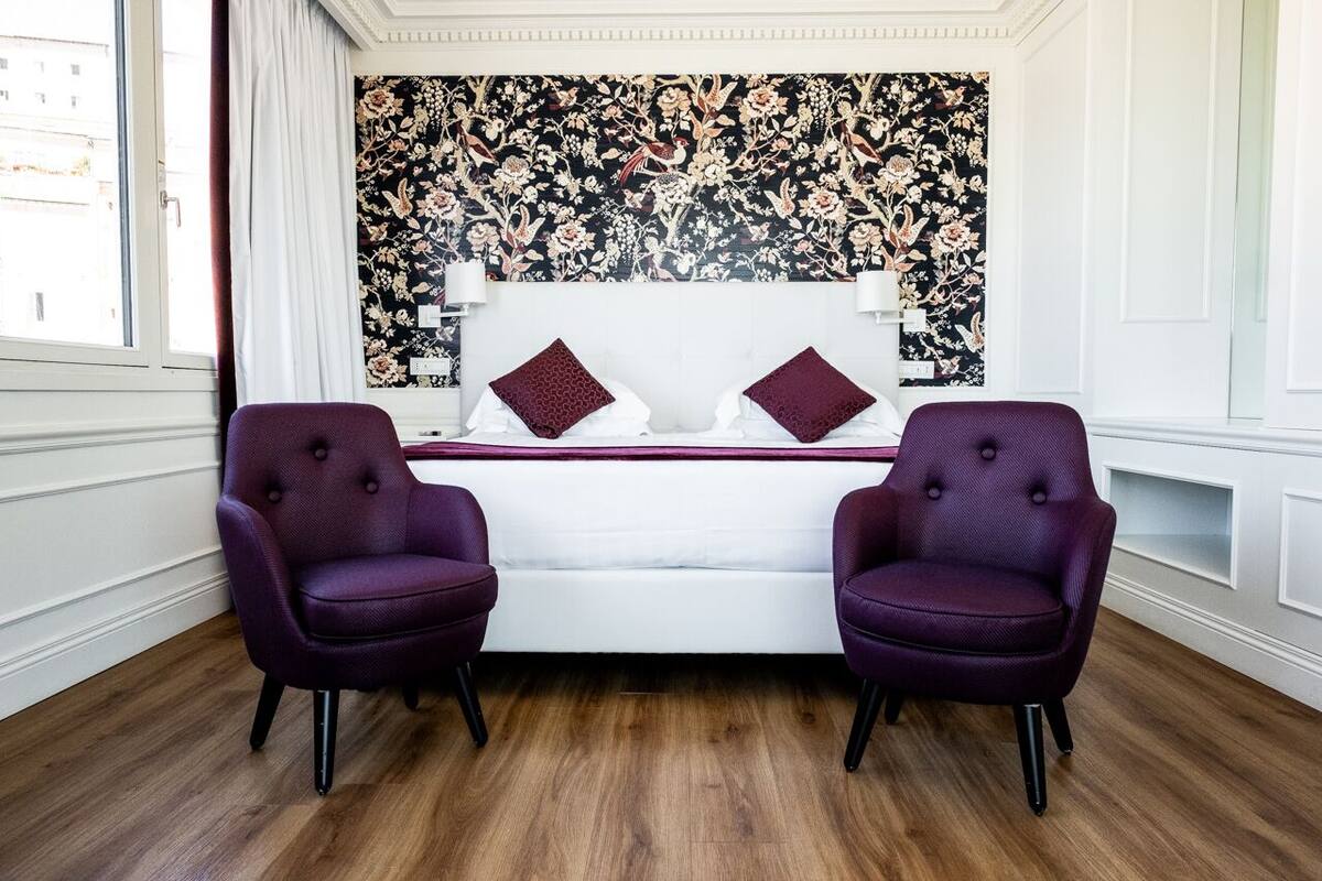 A bedroom with a white bed, two purple armchairs, a floral wallpaper, and a wooden floor.