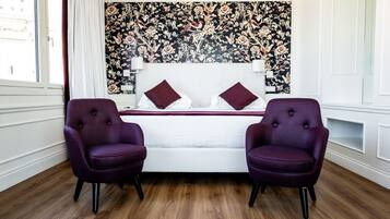 Un dormitorio con una cama blanca, dos sillones morados, un papel tapiz floral y suelo de madera.