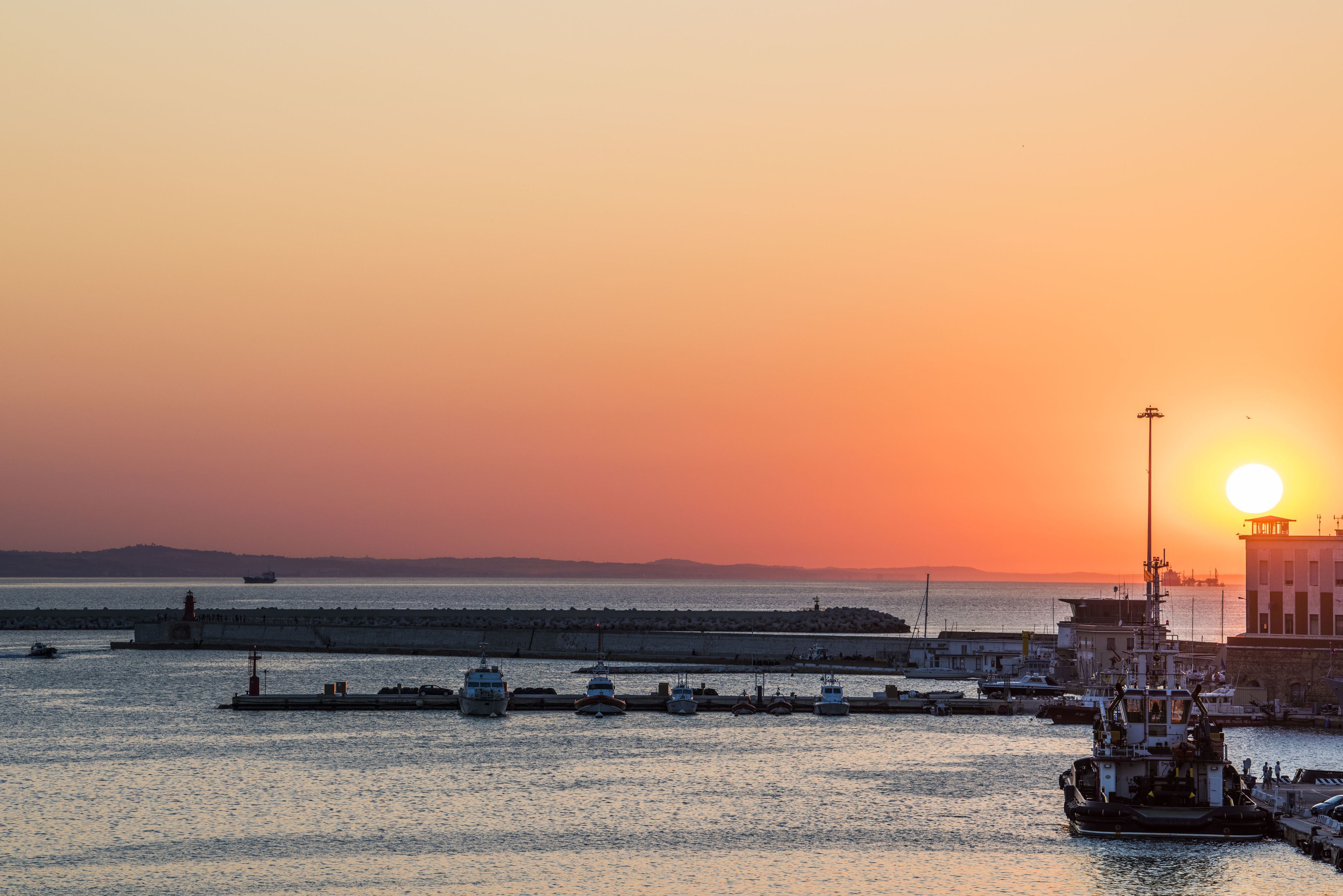 A harbor at sunset with boats docked, a lighthouse, and a building with a flagpole.