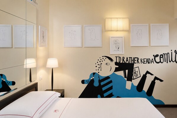 Una habitación de hotel con una cama, un escritorio, una lámpara y una pared decorada con dibujos enmarcados y un cartel grande.