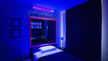 Una camera d'albergo con un letto grande, un comodino con una lampada, una TV a muro e uno specchio con insegna luminosa.