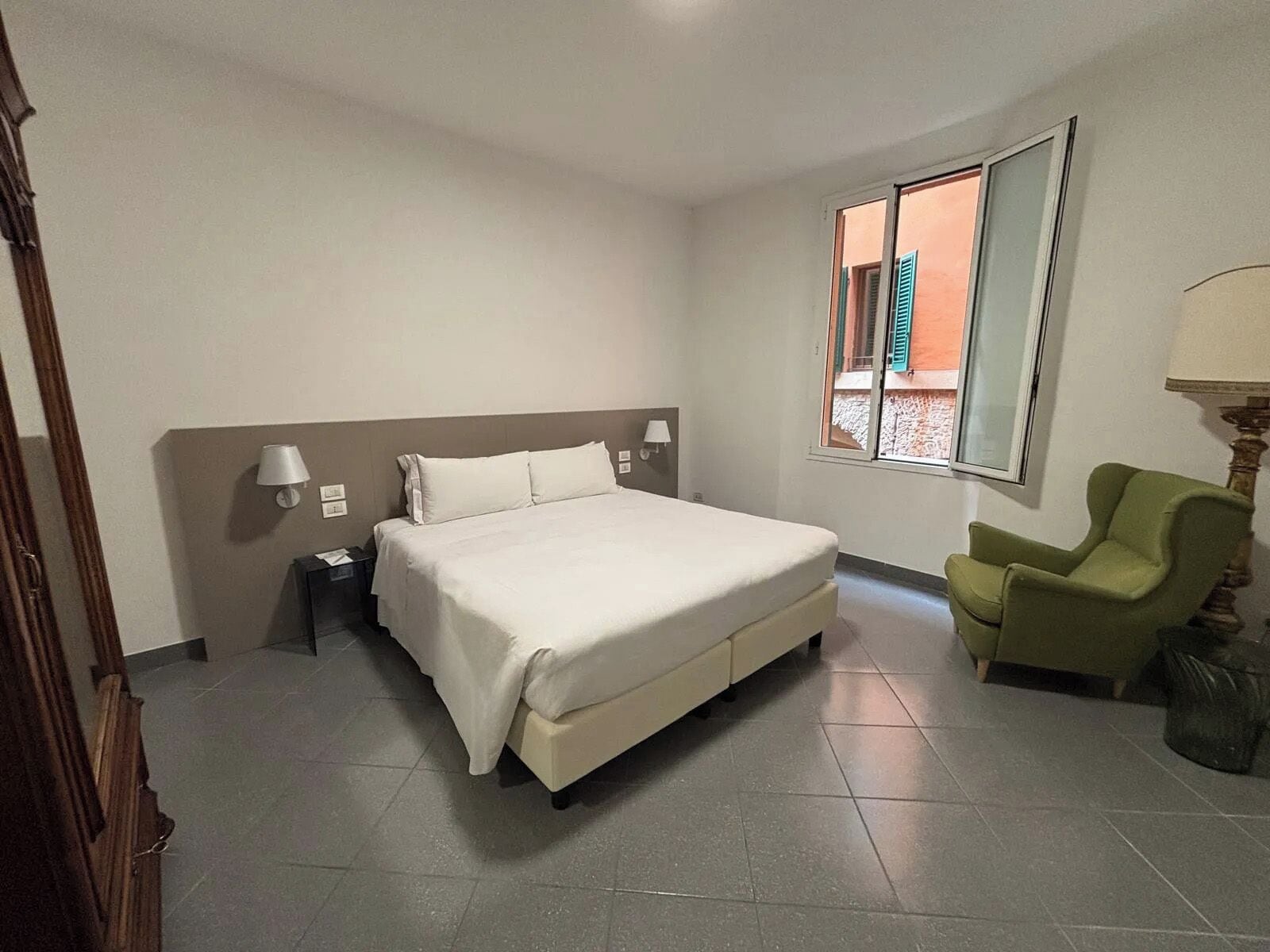 Habitación de hotel con una cama grande, una silla, una lámpara y un ventanal con vistas a edificios.