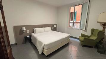 Habitación de hotel con una cama grande, una silla, una lámpara y un ventanal con vistas a edificios.