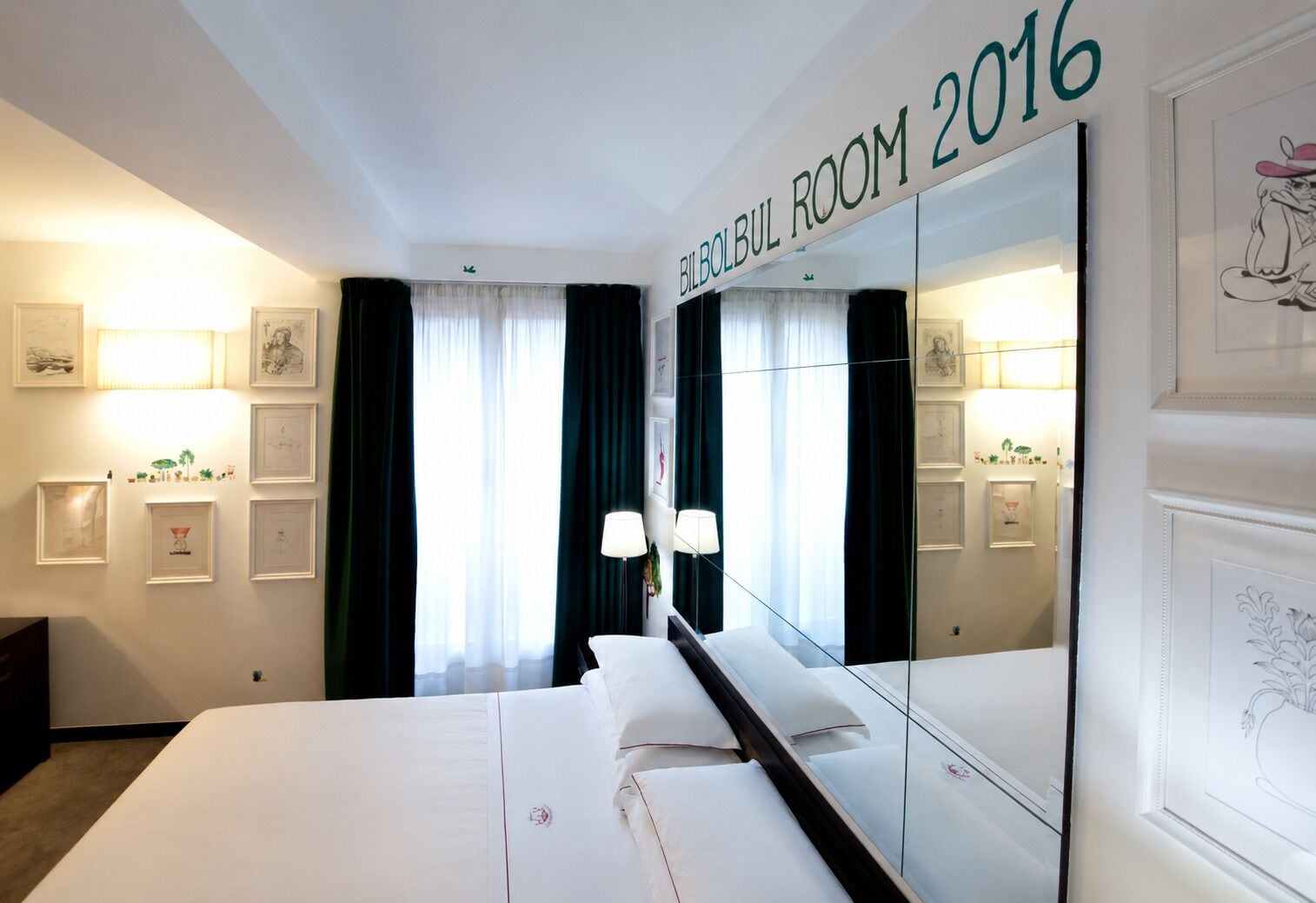 Una camera d'albergo con uno specchio grande, un letto con lenzuola bianche, un comodino con lampade e opere d'arte incorniciate alle pareti.
