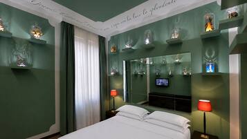 Quarto de hotel com cama, duas mesas de cabeceira, televisão e objetos decorativos nas prateleiras.