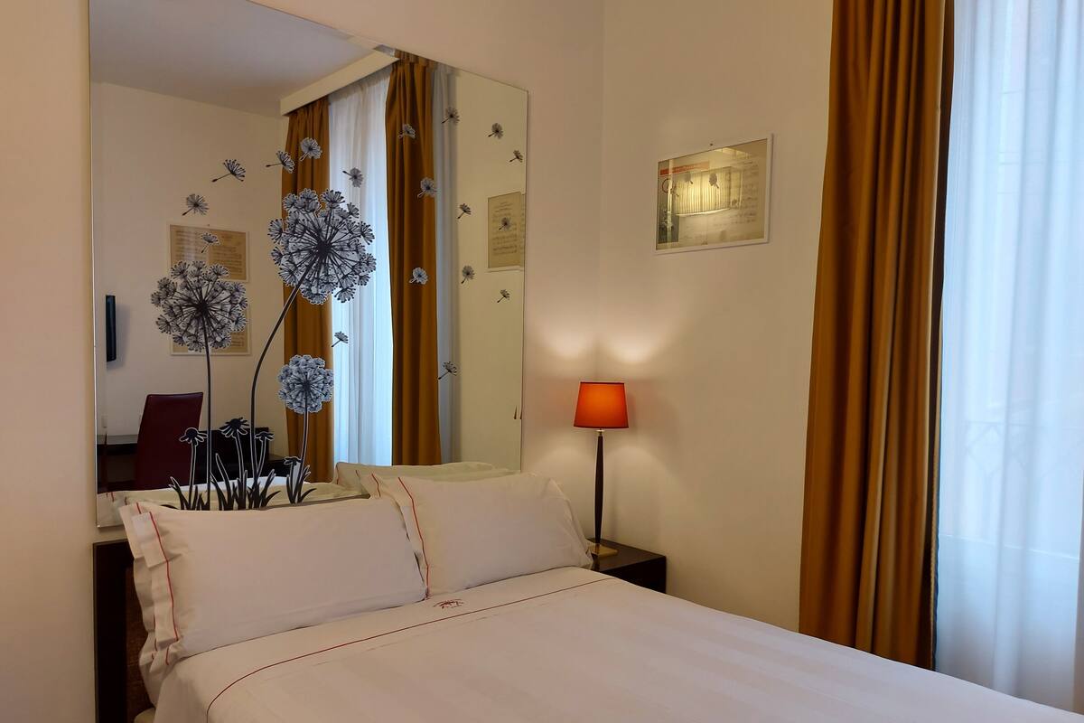 Une chambre d’hôtel avec un lit, une table de chevet avec une lampe, un miroir et une fenêtre avec des rideaux.