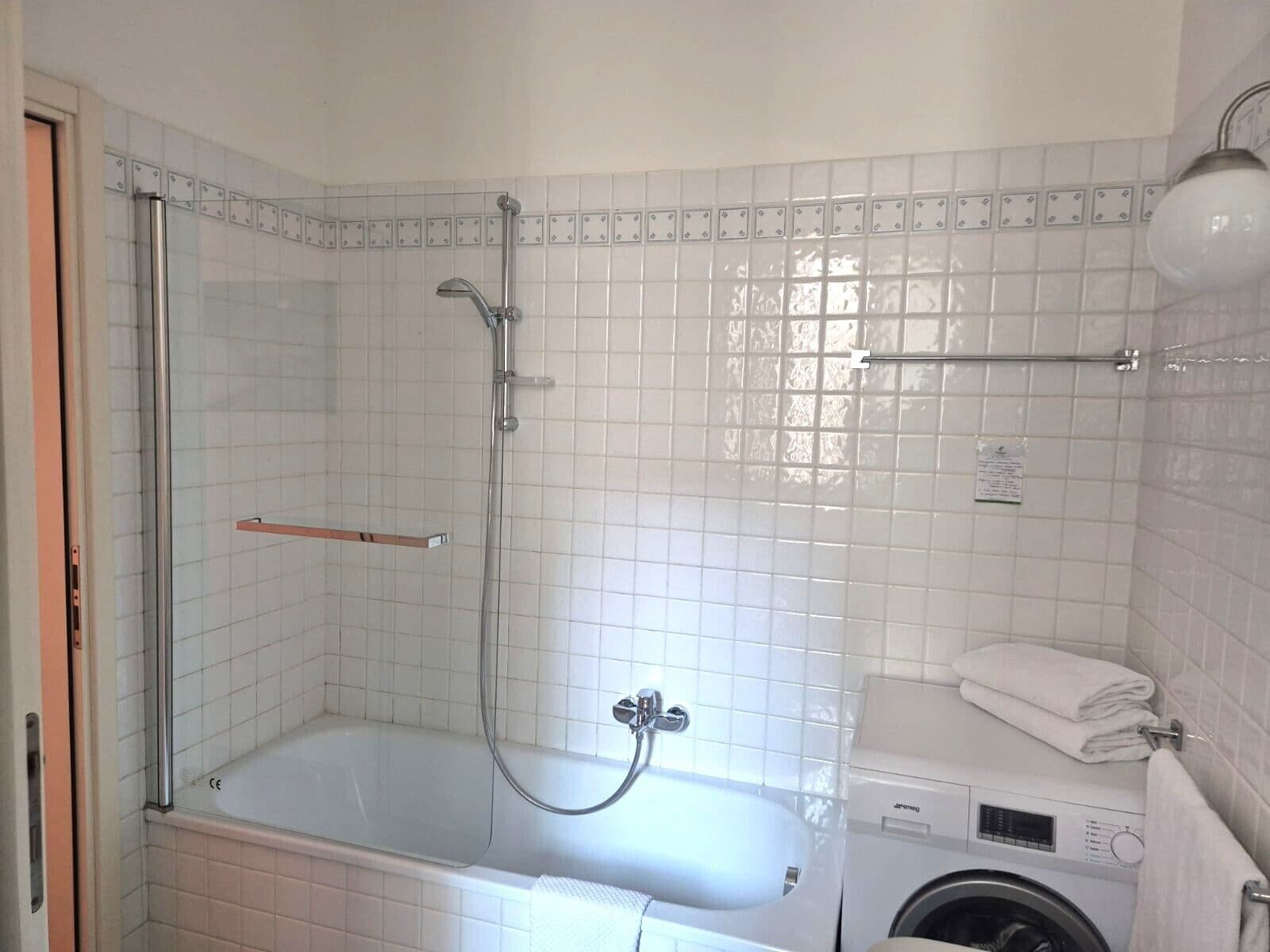 A bathroom with a glass-enclosed shower, a white bathtub, a washing machine, and a towel rack.