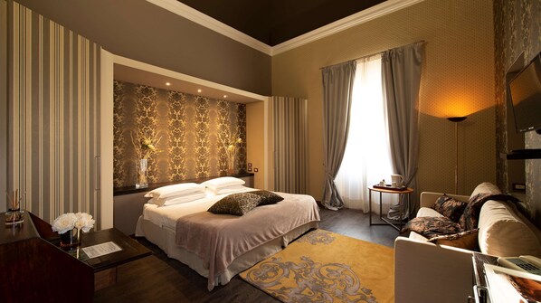 A well-appointed hotel room with a large bed, a sitting area with a sofa and armchairs, a desk with a telephone, and a large window with curtains.