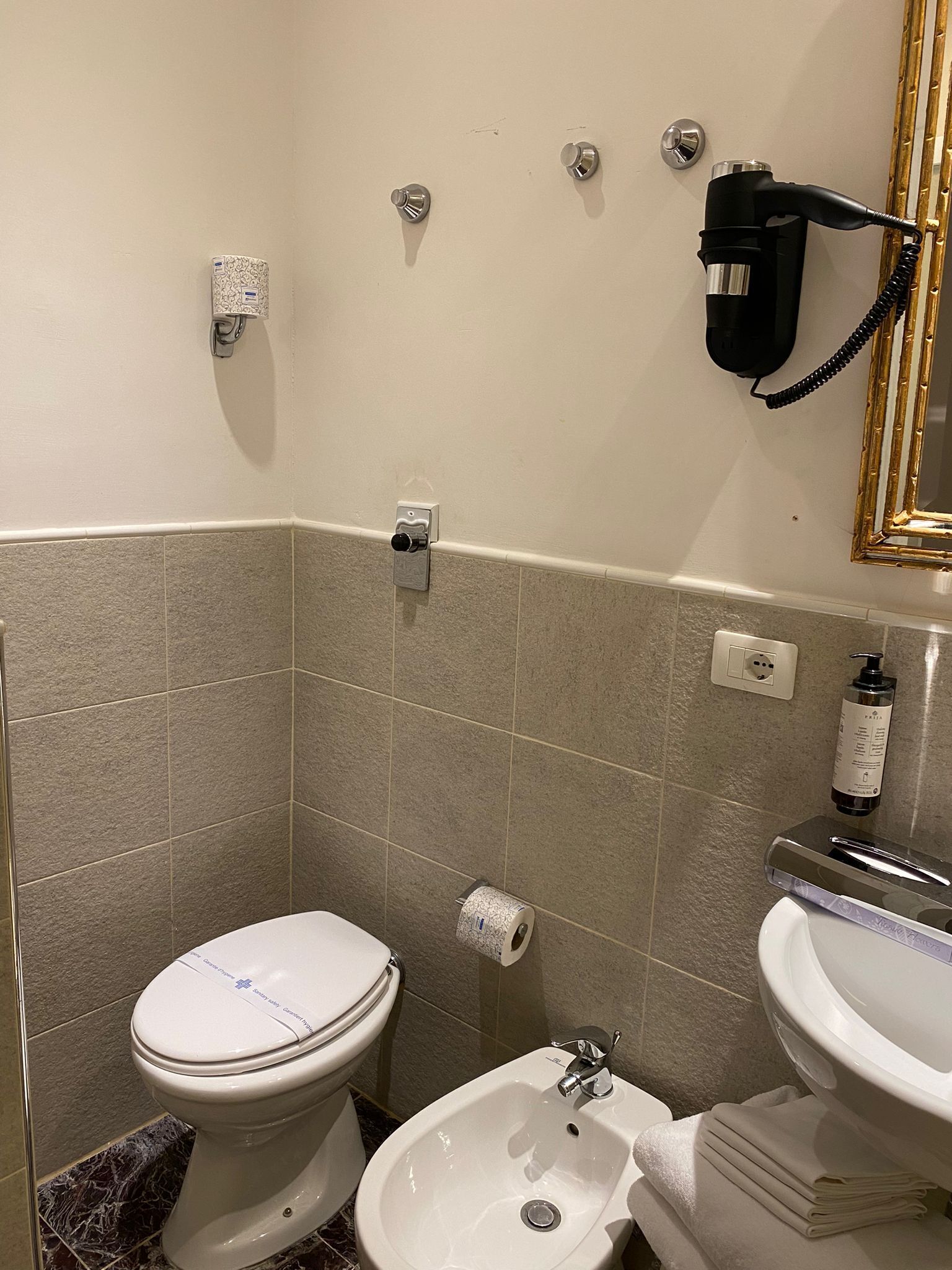 A small bathroom with a toilet, bidet, and sink. There is a hairdryer on the wall and a bottle of hand soap on the sink.