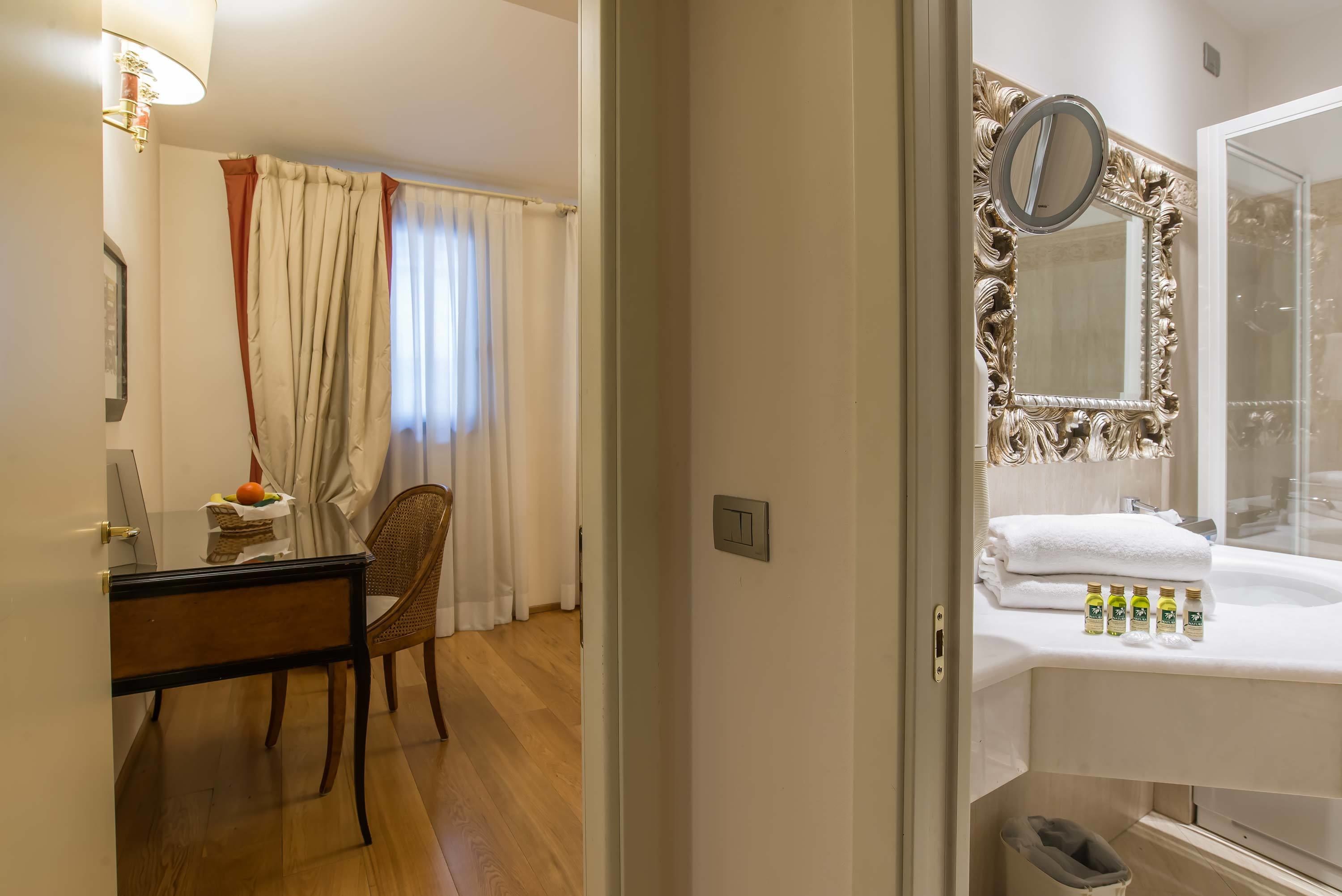 Una camera d'albergo con una scrivania in legno, una sedia e un bagno dotato di uno specchio grande e un lavandino bianco.