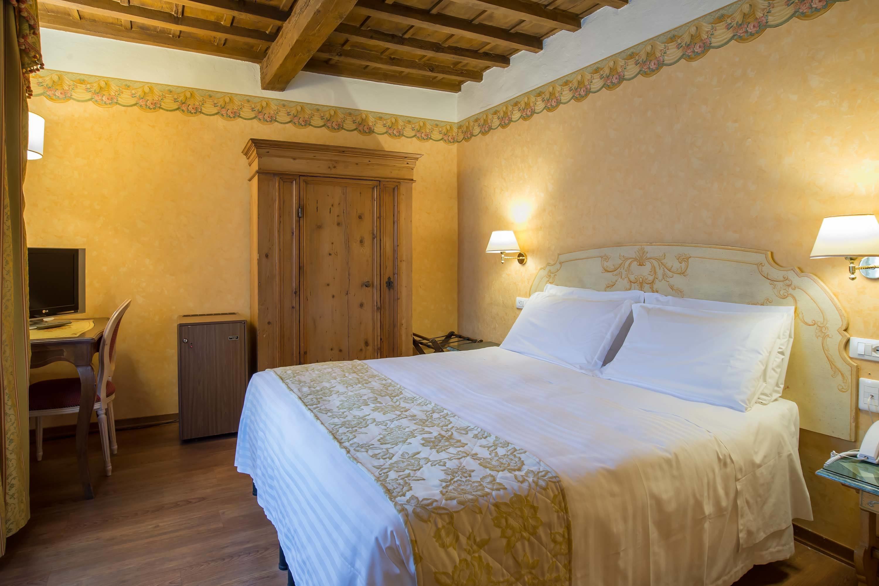 A hotel room with a wooden ceiling, a bed with white linens, a desk with a chair, and a small cabinet.