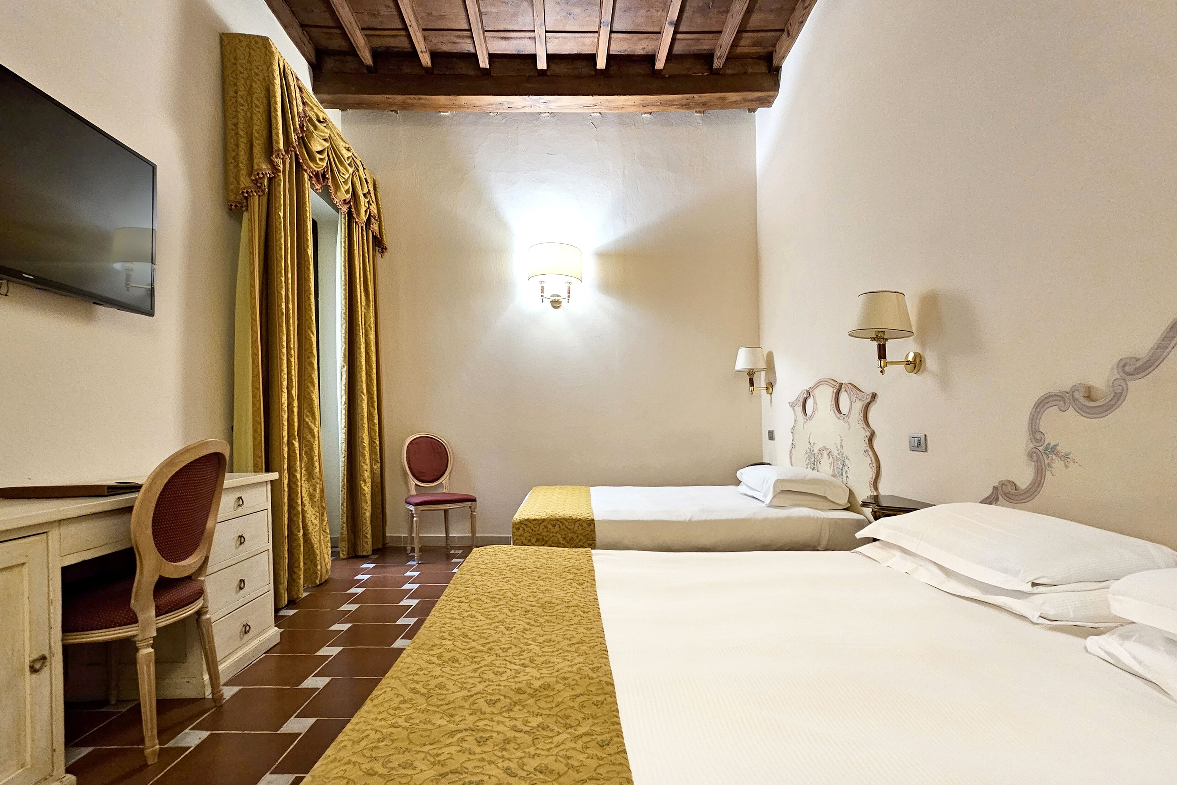 A hotel room with two beds, a desk with drawers, a chair, a television, and a chandelier.