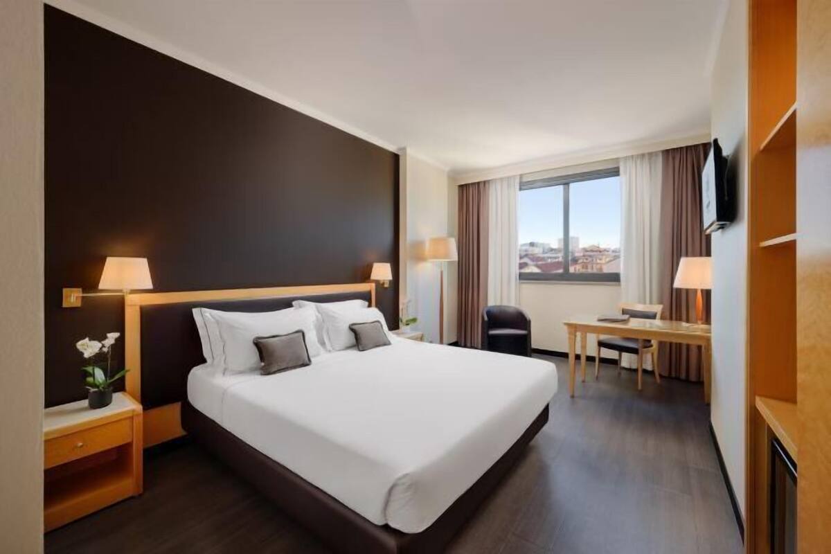 Une chambre d’hôtel avec un grand lit, des tables de chevet avec des lampes, un bureau avec une chaise et une vue sur la ville.