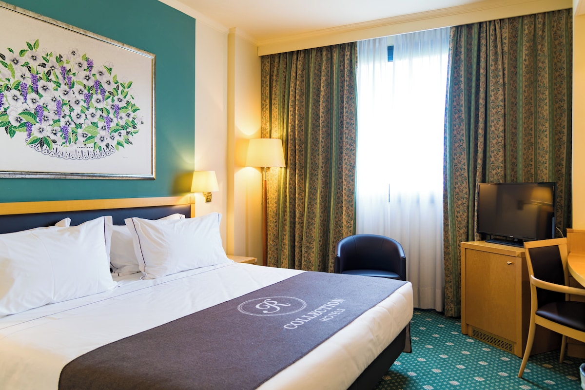 Une chambre d’hôtel comprenant un lit, un bureau, une chaise, un téléviseur et une œuvre d’art florale encadrée accrochée au mur.