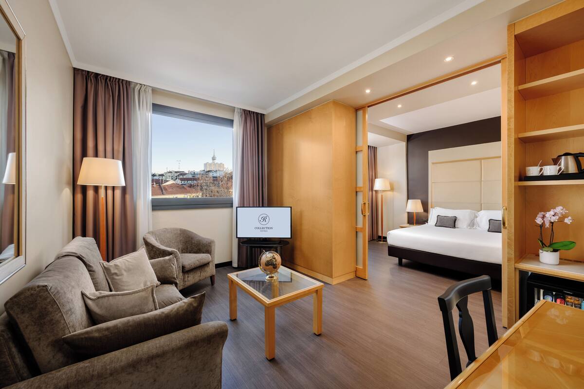 Ein Hotelzimmer mit Sofa, Ohrensessel, Glastisch, Fernseher, Bett, Nachttisch, Lampe und Blick auf die Stadt.