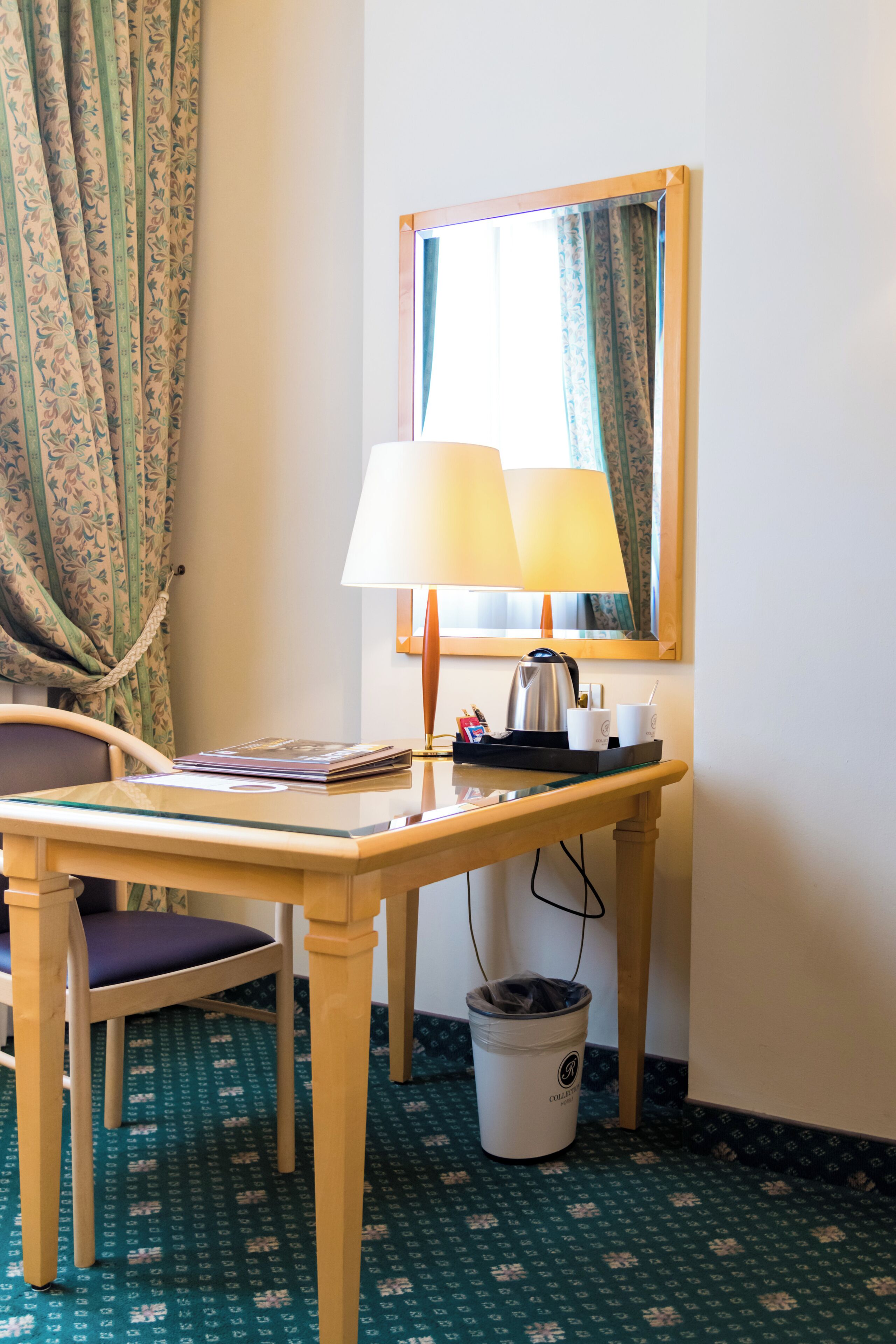 A hotel room with a wooden desk, a chair, a lamp, a coffee maker, a cup, a tray, and a trash bin.
