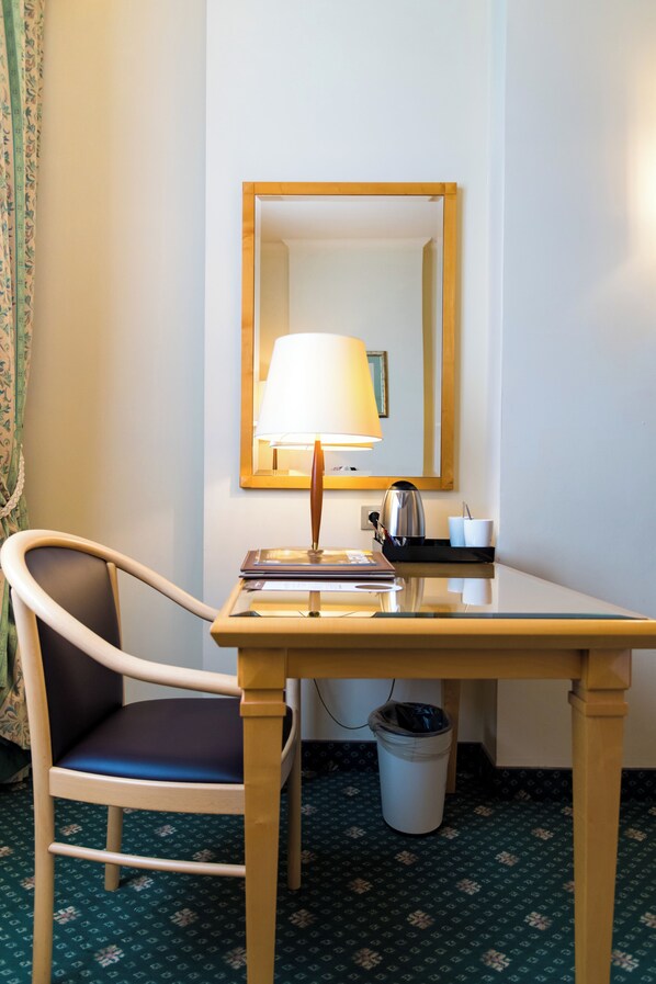 A hotel room with a wooden desk, a chair, a lamp, a trash bin, and a mirror on the wall.