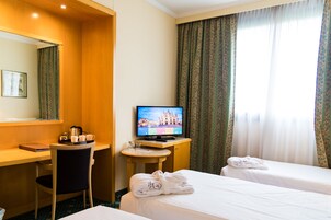 Una camera d'albergo con due letti, una scrivania con televisore, una sedia e una finestra con tende.