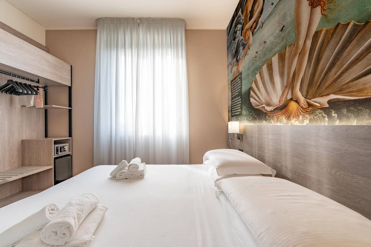 A hotel room with a bed, a wall-mounted TV, a painting, and a window with curtains.