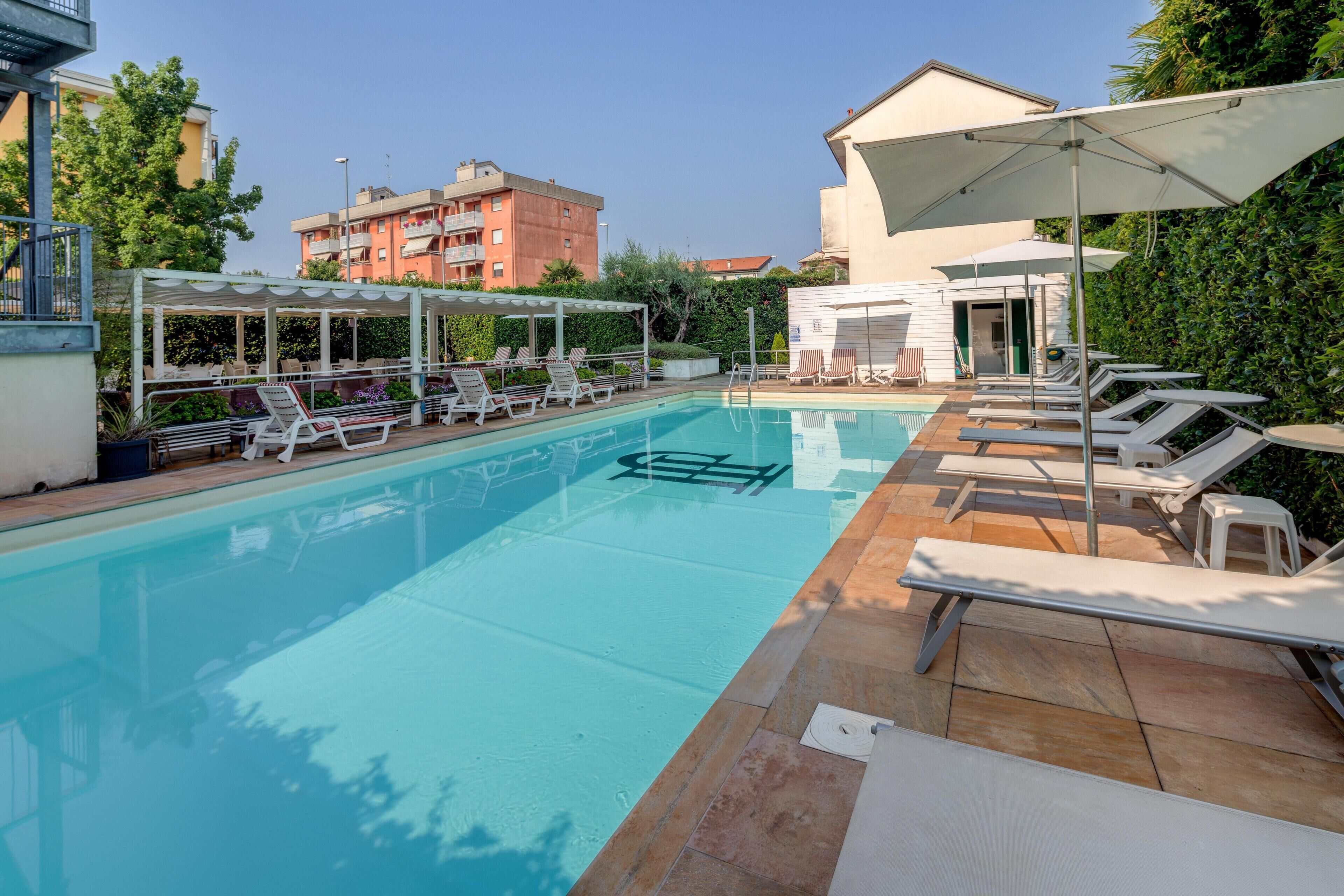 A swimming pool with lounge chairs and umbrellas, surrounded by buildings and greenery.
