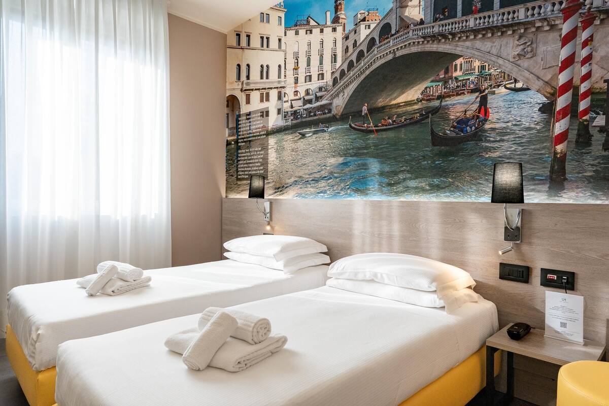 A hotel room with two beds, a mural of a canal scene, a nightstand, and a yellow chair.