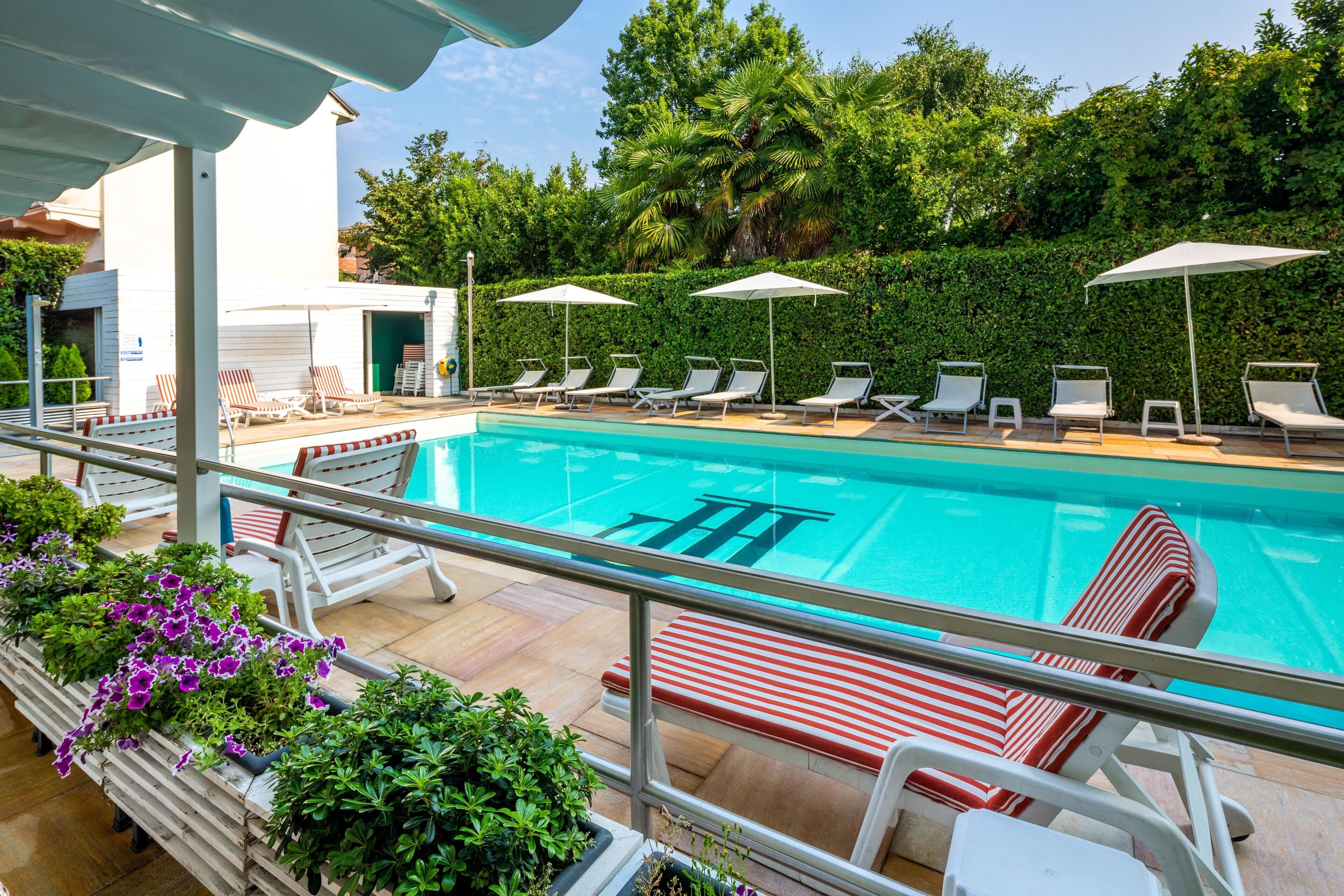 Piscina con tumbonas y sombrillas, rodeada de vegetación y un edificio blanco.