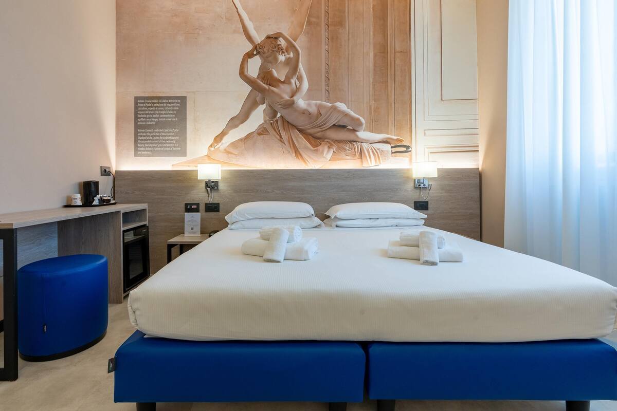 A hotel room with a large bed, a massage table, a desk, and a wall-mounted sculpture.