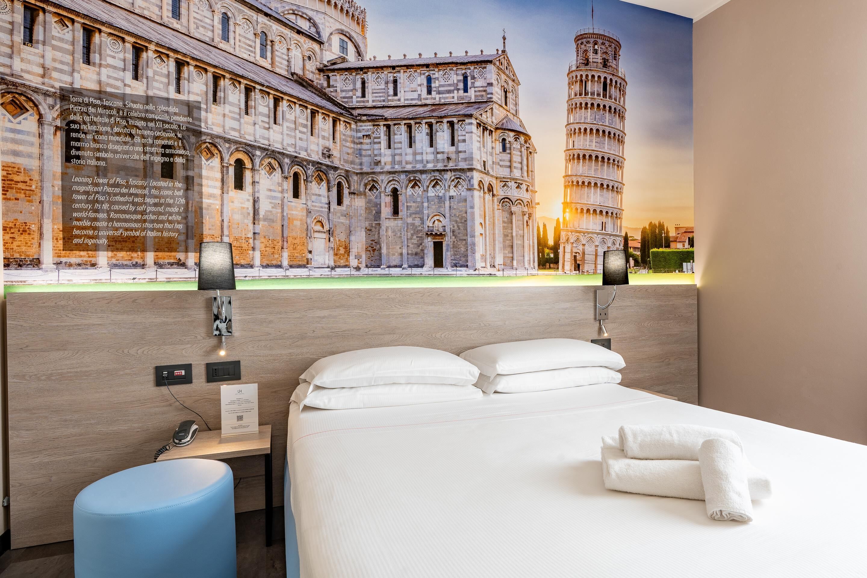 A hotel room with a large bed, a desk with a phone and a lamp, and a mural of the Leaning Tower of Pisa.