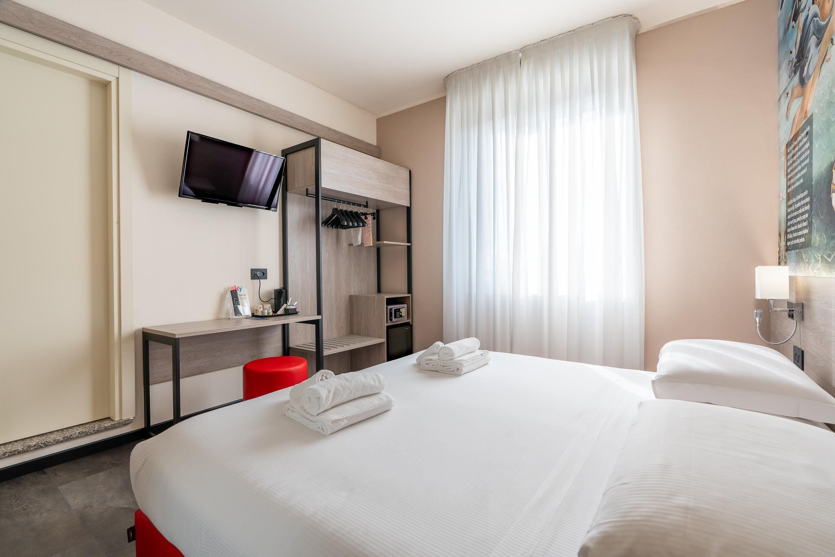 Habitación de hotel con cama, televisor montado en la pared, escritorio con silla y ventana con cortinas.