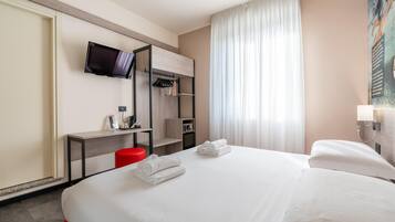 Habitación de hotel con cama, televisor montado en la pared, escritorio con silla y ventana con cortinas.