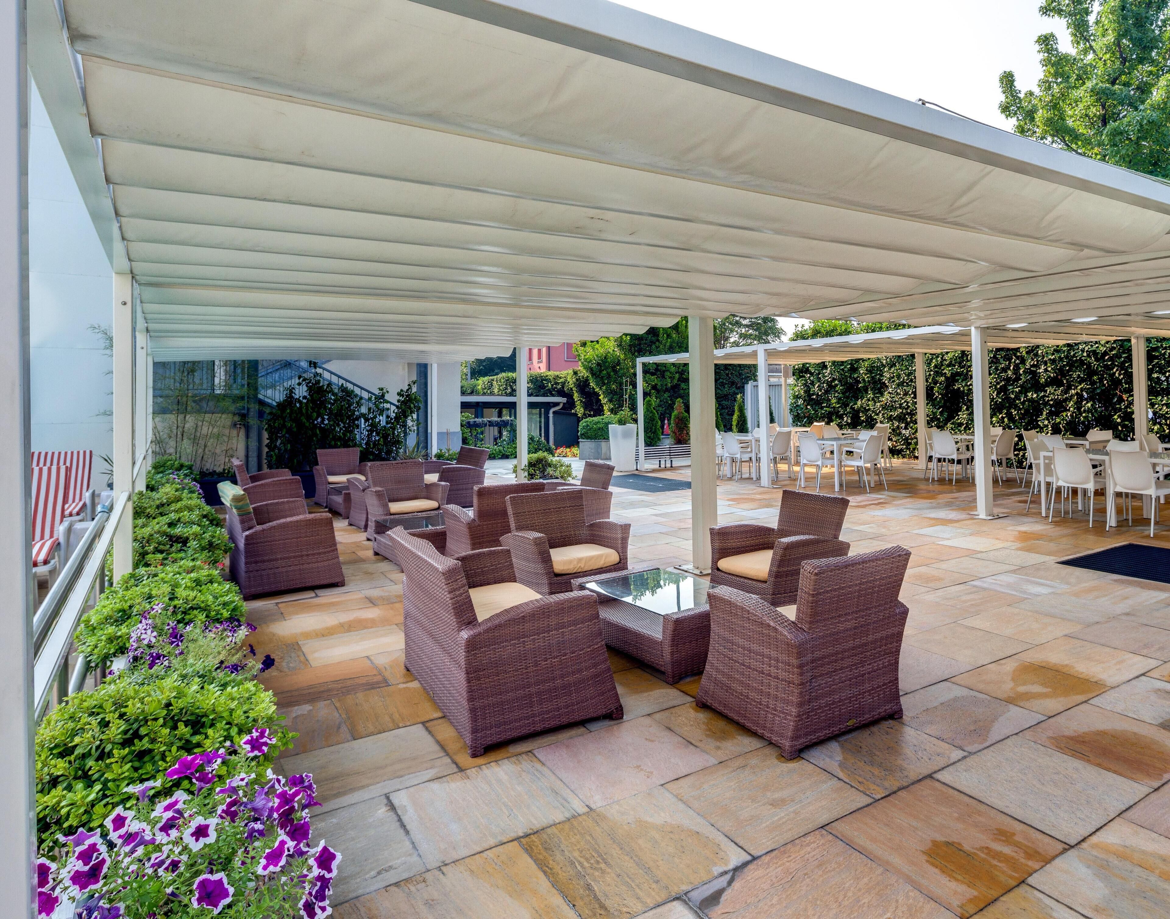 An outdoor patio area with wicker furniture and a glass table, surrounded by stone tiles and covered by a white canopy.